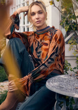 Load image into Gallery viewer, Woman sitting outdoors wearing a leaf patterned blouse spruce green denim and camel-colored boots.