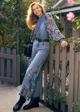 Load image into Gallery viewer, Woman in denim shacket with floral sleeves, jeans with floral side inserts and black boots standing by a wooden fence with a house and pink flowers in the background.