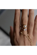 Load image into Gallery viewer, A close-up of a hand wearing a gold wrap ring with a pearl and moonstone and holding a gold chain against a blurred gray background.