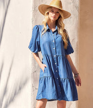 Load image into Gallery viewer, Woman wearing a blue chambray dress and straw hat, with her hand in a side pocket, against a light-colored wall.