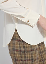 Load image into Gallery viewer, Close-up of a cream-colored blouse with bronze embellishments on the sleeve and cuffs and plaid pants against a plain background.