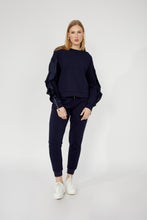 Load image into Gallery viewer, Woman wearing a navy blue ruffled sleeve with zippers top, navy blue pants and white sneakers against a white background.