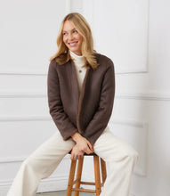 Load image into Gallery viewer, Woman wearing a brown reversible jacket over a white top and pants, sitting on a wooden stool against a white wall.