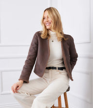 Load image into Gallery viewer, Woman wearing a brown reversible jacket over a white top and pants, sitting on a wooden stool against a white wall.