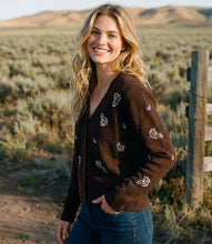 Load image into Gallery viewer, A woman stands outdoors in a desert landscape wearing a dark brown knit cardigan with rhinestone floral embroidery and blue jeans.
