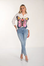 Load image into Gallery viewer, Woman wearing a white blouse with a colorful butterfly design, light blue jeans with embellished hem and silver heels on a white background.