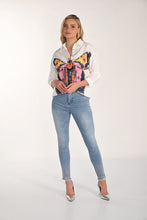 Load image into Gallery viewer, Woman wearing a white blouse with a colorful butterfly design, light blue jeans with embellished hem and silver heels on a white background.