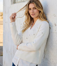 Load image into Gallery viewer, A woman with blonde hair leans against a white textured wall, wearing a cream-colored knit layered sweater with rhinestone embroidered floral details.