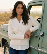 Load image into Gallery viewer, A woman with long dark hair leans against the open door of a vintage sage green truck outdoors. She is wearing a cream-colored layered knit sweater with rhinestone floral applique and a V-neckline, and paired with dark blue jeans.