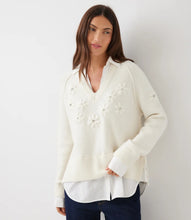 Load image into Gallery viewer, A woman wearing a cream-colored layered knit sweater with white floral appliques and rhinestone details and dark blue jeans, against a plain light gray background.