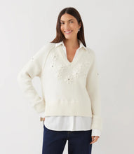 Load image into Gallery viewer, A woman modeling a cream layered knit sweater with 3D floral applique on the chest and dark blue jeans against a plain white studio background.
