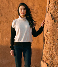 Load image into Gallery viewer, A woman wearing a colorblock turtleneck top with a cream body and black sleeves, paired with dark blue jeans, standing against a textured brown adobe wall.