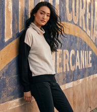 Load image into Gallery viewer, A woman leans against a weathered blue and tan wall, wearing a color-block ribbed knit turtleneck top with a cream body and black sleeves, paired with black pants.