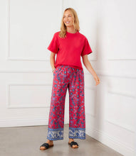 Load image into Gallery viewer, Woman wearing a red short sleeve top and patterned pants against a white wall.