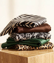 Load image into Gallery viewer, A stack of folded scarves displayed on a light wood block against a warm cream background. The fabrics include zebra print, brown, black and white paisley, dark green, and leopard print.