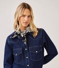 Load image into Gallery viewer, A woman wearing a dark indigo denim western-style shirt jacket with silver snap buttons and chest pockets, styled with a black and white patterned neck scarf, against a light gray studio background.