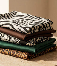 Load image into Gallery viewer, A stack of folded scarves displayed on a light wood block against a warm cream and brown background. The fabrics include zebra print, brown, black and white paisley, dark green, and leopard print.
