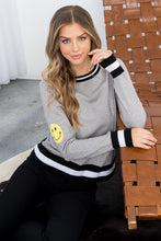 Load image into Gallery viewer, The Heather Gray Smiley Face Knit Sweater - TMK1517 from THML features classic black and white stripes on a gray background but adds a cheerful touch with a darling yellow smiley face on the upper part of each sleeve. This lightweight sweater has a vintage feel and modern appeal, with striping at the bottom hem, cuffs, and neck. Color- Gray, black, white and yellow. Smiley face on upper sleeves. Black and white striping on neckline, sleeves and bottom hem. Lightweight sweater.