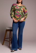 Load image into Gallery viewer, Woman wearing a floral top, blue jeans and metallic flats against a plain background with a barstool in the background.