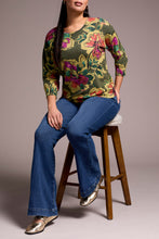 Load image into Gallery viewer, Woman wearing a floral top, blue jeans and metallic flats against a plain background with a barstool in the background.