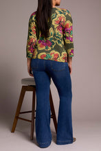 Load image into Gallery viewer, Woman wearing a floral top, blue jeans and metallic flats against a plain background with a barstool in the background.