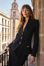 Load image into Gallery viewer, Woman wearing an indigo fitted denim jacket with gold buttons and denim skirt standing on a balcony.