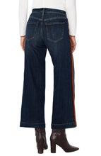 Load image into Gallery viewer, Model wearing dark blue wide leg jean with brown multicolor striping on sides, white long sleeve top and brown boots against a white background.