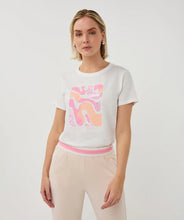 Load image into Gallery viewer, Woman wearing a white t-shirt with pink graphic design and tan pants on a plain background.