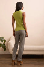 Load image into Gallery viewer, Woman wearing a green sleeveless top, gray pants and brown boots standing in a room with a beige sofa and plant.