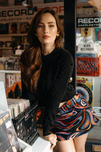Load image into Gallery viewer, Woman in a record store with vinyl records and shelves in the background, wearing a black sweater and abstract print mini skort.