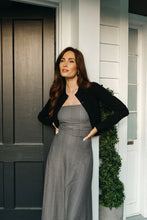 Load image into Gallery viewer, Woman wearing a long checkered dress and black cardigan standing in front of a house with a wreath on the door.