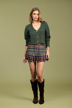 Load image into Gallery viewer, Woman wearing a green cardigan, multi-color plaid skirt and brown boots against a pale green background.
