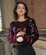 Load image into Gallery viewer, Woman wearing a sequined maroon flared sleeve blouse and black pants standing against a brick wall.