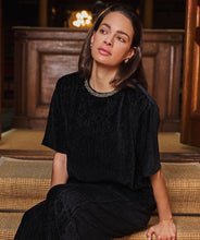Load image into Gallery viewer, Woman wearing a black velvet short sleeve top with embellished neckline and black velvet pants sitting on a wooden floor in a warm, indoor setting.