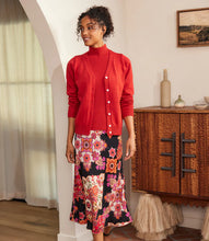 Load image into Gallery viewer, Woman wearing a red cardigan over a coordinating turtleneck and printed skirt, standing in a room with a wooden cabinet and picture on the wall.