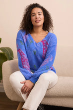 Load image into Gallery viewer, Woman wearing a blue sweater with colorful patterns, off-white pants and black boots sitting on a beige chair.