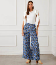 Load image into Gallery viewer, Woman wearing a white top and blue floral pants against a white paneled wall.