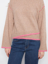 Load image into Gallery viewer, A close-up view of a person wearing a beige knit sweater with bright pink contrast trim along the shoulders, hem, and cuffs, paired with blue jeans against a plain light background.
