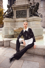 Load image into Gallery viewer, Woman sitting on a stone bench in front of a statue in an urban setting wearing a black and white contrast tunic blouse with jewel embellishments, black pants and black boots.
