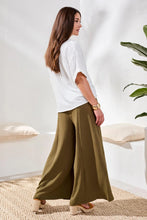 Load image into Gallery viewer, Woman wearing a white blouse and olive green wide-leg pants in a bright room with a plant.
