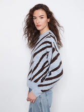 Load image into Gallery viewer, A woman with long, wavy brown hair is shown from the side, wearing a light blue-gray and dark brown zebra-striped knit sweater and light-wash blue jeans against a plain white studio background.
