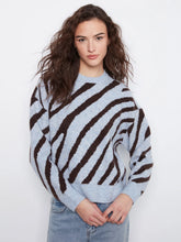 Load image into Gallery viewer, A woman wearing a light blue and dark brown zebra-striped knit sweater with light-wash blue jeans, standing against a plain white studio background.
