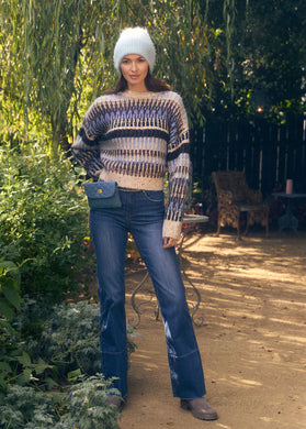 Woman wearing a multi blue and sparkle sweater, blue jeans, grey boots, fuzzy winter cap and carrying a blue handbag standing outside.
