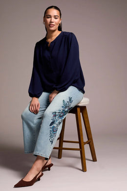 Woman sitting on a stool wearing a navy blue top, ight blue jeans with blue floral embroidery and brown heels.
