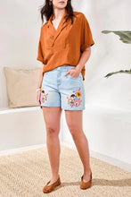 Load image into Gallery viewer, Woman wearing a rust-colored top, light blue shorts with floral embroidery and brown flats in a room with a plant and pillow in background.
