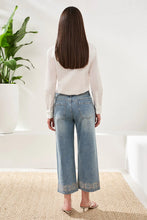 Load image into Gallery viewer, Woman wearing a white blouse, blue jeans with embellishments and sandals standing in a room with a plant and white wall.

