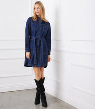 Load image into Gallery viewer, Woman wearing a belted blue denim dress and tall black suede boots against a white wall.
