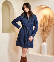 Load image into Gallery viewer, Woman wearing a belted blue denim dress and tall brown boots standing in a room with decorative elements.
