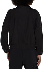 Load image into Gallery viewer, The Brylee utility jacket from Liverpool Los Angeles features a cropped design and cinched hem, perfect for adding a touch of edginess to any outfit. Made with a textured stretch woven fabric, this jacket offers both style and comfort. Stay effortlessly stylish with this versatile piece.  Color- Black. Collared. Hidden button placket with zip front closure. Two front flap pockets with double button closure. Button tab at arm to roll sleeves. Cinch waist. Cropped.
