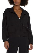 Load image into Gallery viewer, The Brylee utility jacket from Liverpool Los Angeles features a cropped design and cinched hem, perfect for adding a touch of edginess to any outfit. Made with a textured stretch woven fabric, this jacket offers both style and comfort. Stay effortlessly stylish with this versatile piece.  Color- Black. Collared. Hidden button placket with zip front closure. Two front flap pockets with double button closure. Button tab at arm to roll sleeves. Cinch waist. Cropped.
